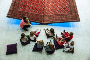 A Teacher kneeling in front of students sitting on the floor